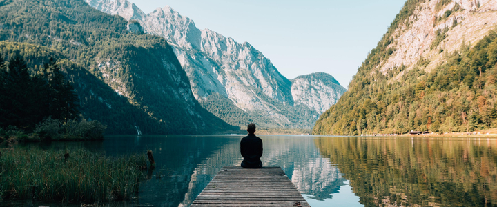 A man sat at the end of a jetty over a lake surrounded by high mountains