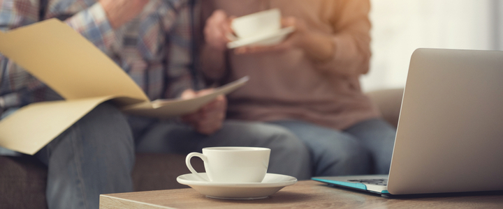 A mug and a laptop on a coffee table with two elderly people behind, looking at paperwork